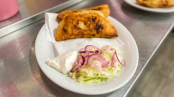 Two golden-brown empanadas with a side of shredded lettuce, onions, cilantro, and a dollop of sour cream on a red plate.