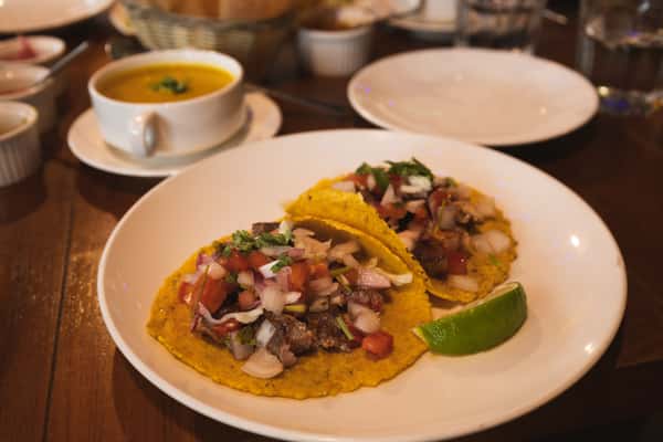 Street-style tacos topped with grilled meat, fresh pico de gallo, diced onions, and cilantro, served on corn tortillas with a lime wedge and soup on the side.