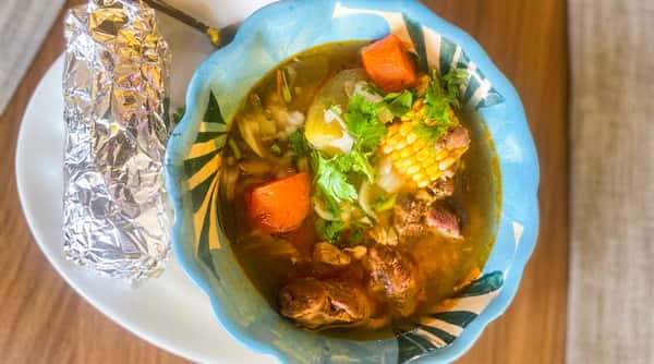 Hearty soup with chunks of meat, corn on the cob, carrots, onions, and cilantro in a blue bowl.