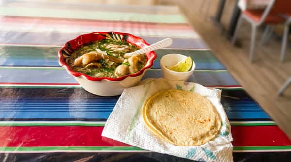 Menudo soup in a red and white bowl with spoon, served with tortillas and a lime wedge.