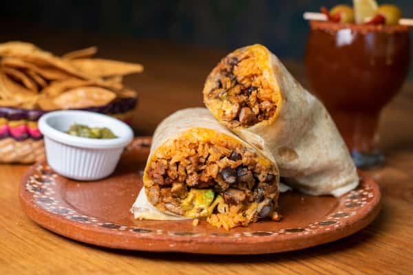 Cut burrito filled with seasoned meat, Mexican rice, beans, and avocado on a rustic ceramic plate, served with salsa, tortilla chips, and a michelada-style cocktail.