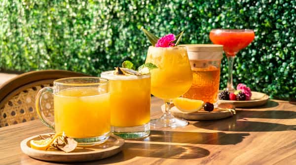 A variety of cocktails, including orange and pink beverages, on a wooden table with a green hedge background.