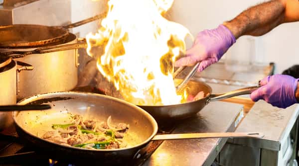Chef in purple gloves stir-frying food in a pan with large flames in a commercial kitchen.