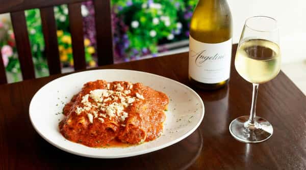 Cannelloni pasta with red sauce and shredded white cheese, a bottle of white wine, and a full glass of white wine on a wooden table.