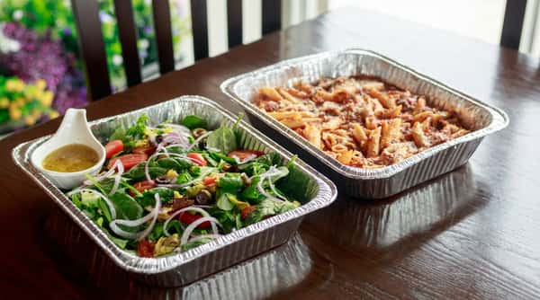 Foil trays of garden salad with dressing and penne pasta with sauce and cheese on a dark wood table.