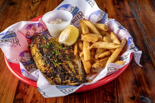 Blackened fish filet garnished with herbs, accompanied by seasoned fries, tartar sauce, and a lemon wedge, served in a branded seafood basket.