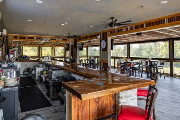 Interior of a rustic, open-air bar and dining area featuring a polished wooden bar, ceiling fans, and scenic views through large screened windows.