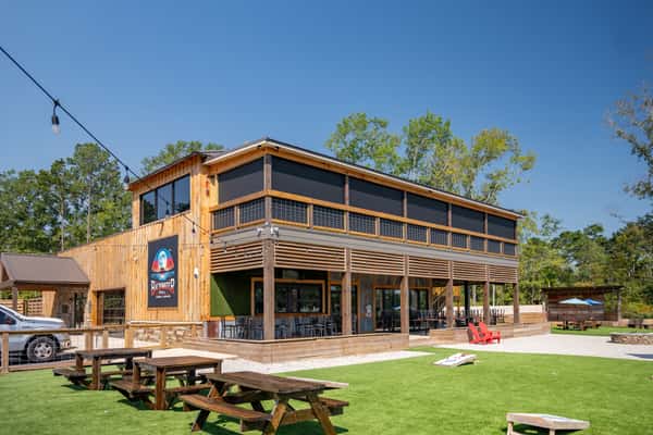 Exterior of Backwater restaurant featuring a modern wooden structure with outdoor picnic seating and green turf, set against a bright blue sky.