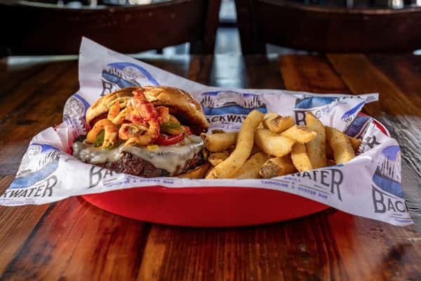Spicy fried shrimp burger topped with jalapeños and melted cheese, paired with thick-cut fries in a red basket, served on branded paper at a coastal-style grill.