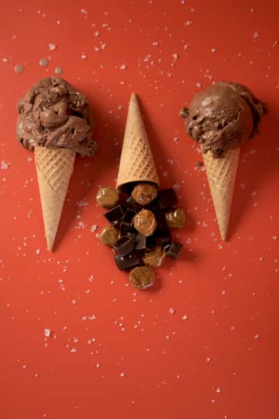 Chocolate ice cream cones with caramel candies and chocolate chunks on a bright orange background, indulgent sweet treat flat lay
