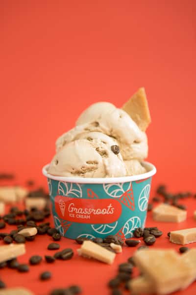 Coffee-flavored ice cream scoops in a branded cup with coffee beans and candy pieces on vibrant orange background, gourmet dessert styling