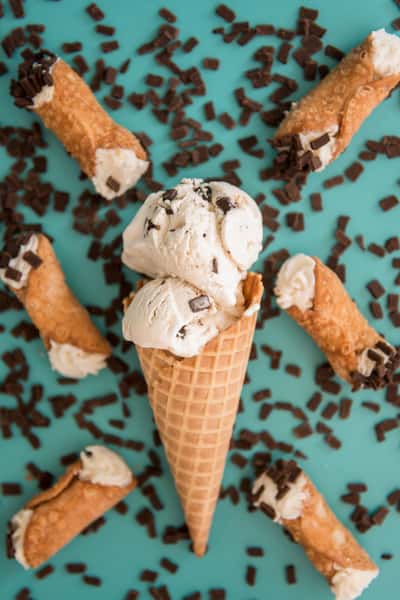 Chocolate chip ice cream in a waffle cone with cannoli shells and chocolate sprinkles on teal background, artisanal dessert flat lay