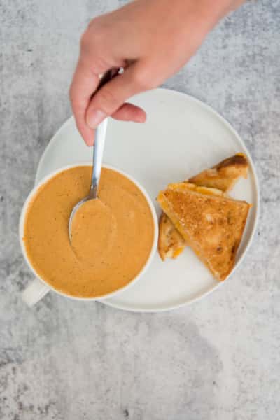 Creamy tomato soup in a white bowl with a grilled cheese sandwich on a plate, hand stirring with spoon, cozy comfort food flat lay
