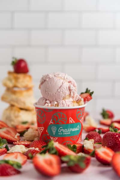 Strawberry ice cream scoop in a branded cup surrounded by fresh strawberries and shortcake pieces, vibrant dessert shop display