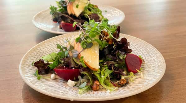 Salad with mixed greens, red beets, sliced apples, crumbled white cheese, and candied nuts on a textured white plate.