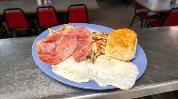 Breakfast plate with two fried eggs, ham, hash browns, and a biscuit on a blue plate.