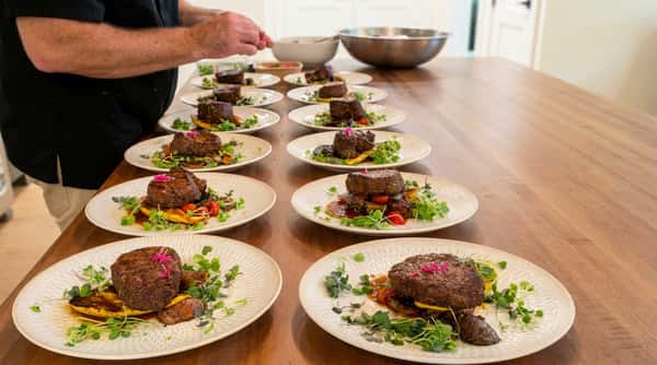 Chef plating multiple dishes of steak with microgreens, roasted vegetables, and sauce on white plates.