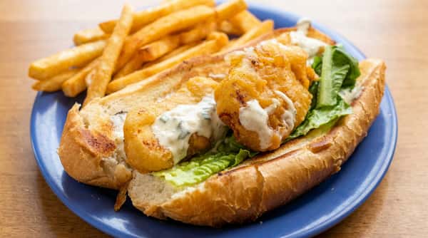 Fried fish po'boy sandwich with lettuce and tartar sauce, served with French fries on a blue plate.