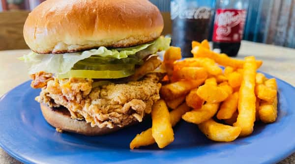 Fried chicken sandwich with lettuce and pickles, served with golden french fries and a soda in the background.
