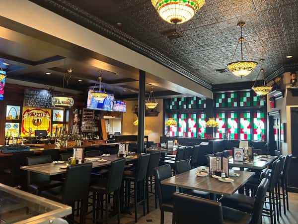 Cozy Italian restaurant bar area with stained glass windows, dark wood furnishings, high-top tables, and vintage-style hanging lights.