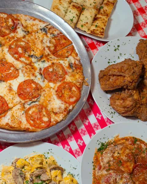 Overhead view of a hearty Italian-American meal spread featuring a tomato and mushroom pizza, garlic breadsticks, crispy fried chicken, tortellini with mushrooms in a creamy sauce, and eggplant parmesan, all served on white plates over a red-and-white checkered tablecloth.