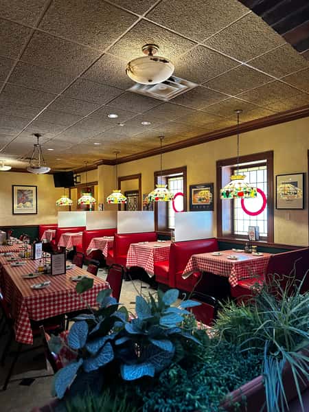 Cozy Italian restaurant interior featuring red leather booths, checkered red-and-white tablecloths, stained glass pendant lights, and framed vintage artwork, creating a warm and nostalgic dining ambiance.