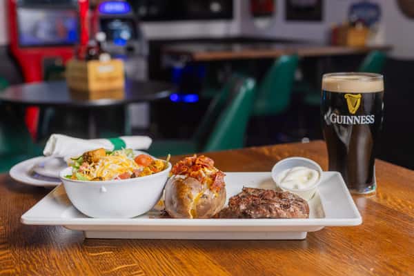 Steak dinner special served with loaded baked potato, coleslaw, and a pint of Guinness in a classic pub setting.