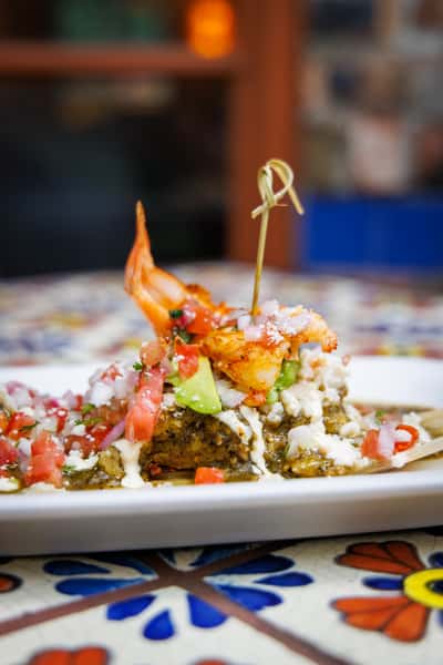 Close-up of a savory tamale topped with grilled shrimp, avocado slices, diced tomatoes, onions, and drizzled crema, served on a corn husk over a traditional Talavera-tiled table.