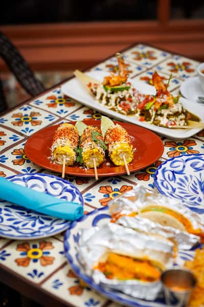 Colorful Mexican street food spread featuring grilled elote (Mexican street corn) on skewers topped with cotija cheese, chili powder, and fresh cilantro, served with lime wedges. Background dishes include tamales with vibrant toppings and tacos wrapped in foil, all set on a vibrant Talavera-tiled table.