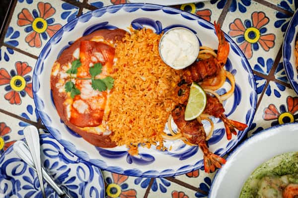 Vibrant Mexican combo plate with cheesy red enchiladas, fluffy Mexican rice, and grilled bacon-wrapped shrimp garnished with a lime wedge and crema, served on a blue-rimmed Talavera-style plate.