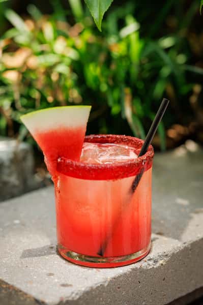 Refreshing watermelon margarita served on the rocks in a glass rimmed with chili salt and garnished with a fresh watermelon slice, set against a lush outdoor backdrop—perfect for summer sipping.