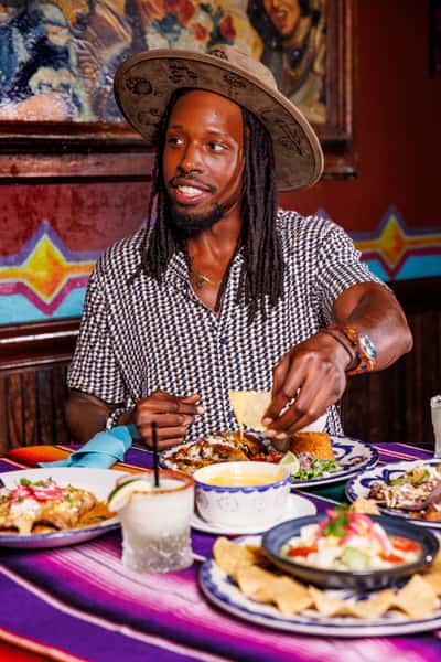 Man enjoying a vibrant Mexican meal at a festive restaurant table, dipping a tortilla chip into creamy queso dip, surrounded by colorful entrées, including enchiladas, ceviche, and margaritas, all set on a striped serape tablecloth.