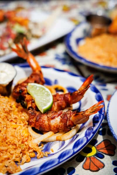 Close-up of bacon-wrapped shrimp and Mexican rice with grilled onions, served on a blue-rimmed Talavera plate with a lime garnish.