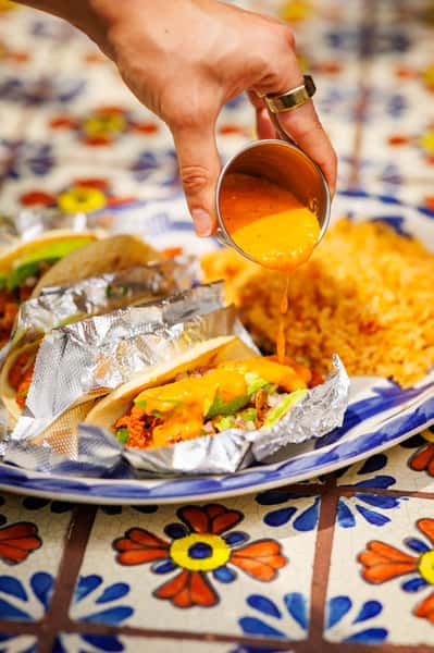 Hand pouring vibrant orange queso sauce over foil-wrapped tacos filled with seasoned meat, fresh onions, and avocado slices, served alongside fluffy Mexican rice on a Talavera-style plate.