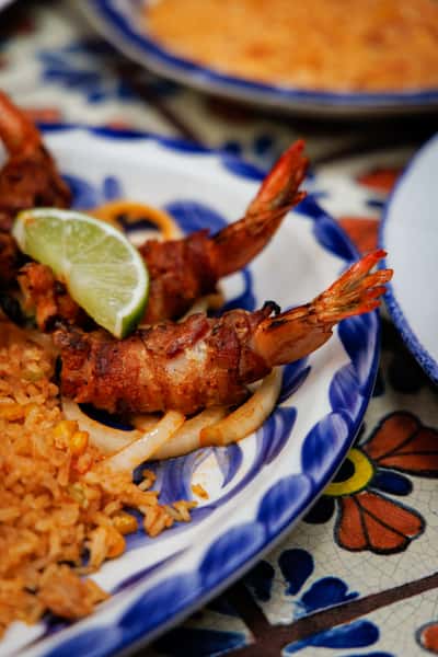Bacon-wrapped shrimp skewers served over grilled onions with a wedge of lime and a side of Mexican rice on a Talavera-style plate.