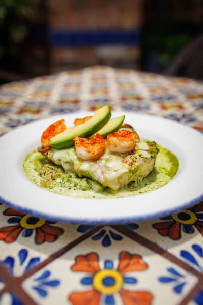 Succulent shrimp and fresh avocado slices layered on top of grilled chicken and creamy cheese, served over a bed of green rice with a poblano cream sauce on a decorative Mexican tile table.