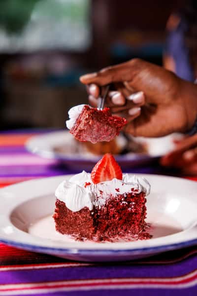 Close-up of a fork lifting a bite of red velvet tres leches cake topped with whipped cream and a fresh strawberry, served in a creamy milk base on a white plate over a festive tablecloth.