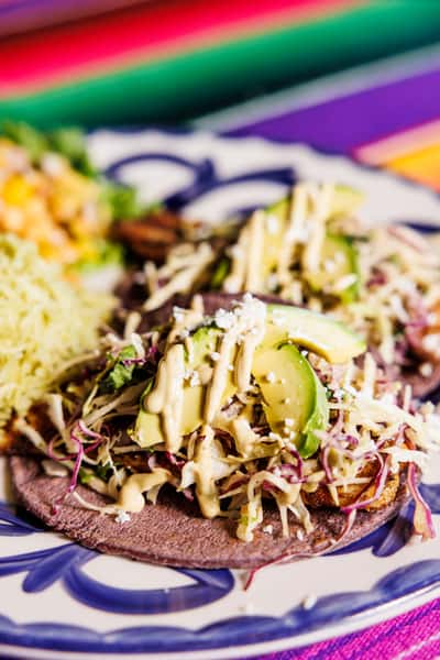 Two blue corn tortillas topped with grilled meat, shredded cabbage, avocado slices, crumbled cheese, and creamy chipotle sauce, served with rice and corn salsa on a traditional Talavera-style plate.