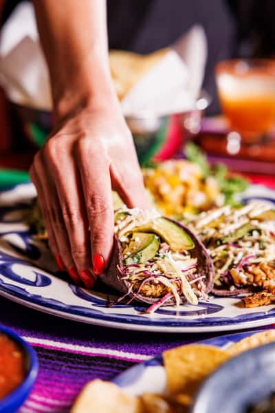 Hand reaching for a blue corn taco filled with grilled protein, slaw, avocado, and queso fresco, plated with rice and corn salad in a festive Mexican restaurant setting.