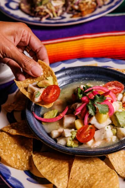 Mexican-style fish ceviche with avocado, cherry tomatoes, red onions, and cilantro in citrus marinade, served with tortilla chips on a Talavera-style plate over a vibrant tablecloth.