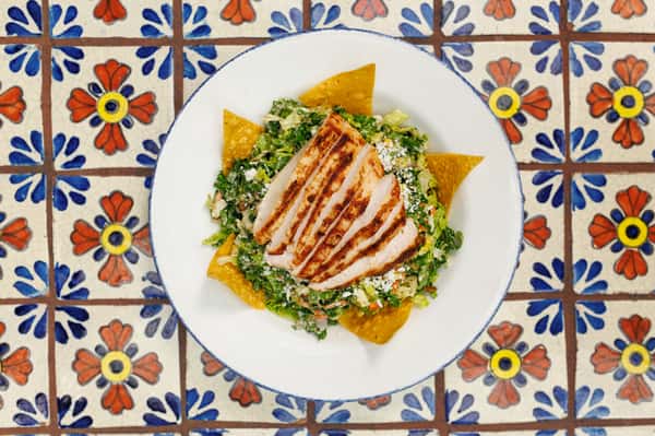 Grilled chicken breast slices atop a bed of chopped romaine, cotija cheese, and tortilla chips, served in a white bowl on a decorative Mexican tile table.