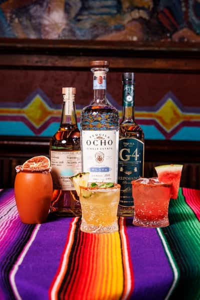 Colorful display of premium tequila bottles—Tequila Ocho, G4, and Casamigos—paired with vibrant cocktails including a clay-rimmed cantarito, pineapple jalapeño margarita, and watermelon margarita, all set on a bold Mexican serape tablecloth in a festive bar setting.