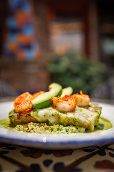 Close-up side view of grilled chicken with shrimp and avocado, smothered in melted cheese and green sauce, presented over rice on vibrant Mexican tilework.