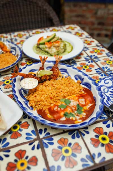 Mexican combo plate featuring enchiladas smothered in red chile sauce, golden Mexican rice, and crispy bacon-wrapped shrimp with lime, served with creamy dipping sauce on a hand-painted blue Talavera plate.