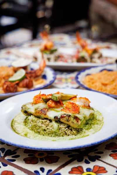Grilled chicken breast topped with shrimp, avocado slices, and melted cheese over cilantro-lime rice, drizzled with creamy green sauce, served on a Talavera-style plate with other Mexican dishes in the background.