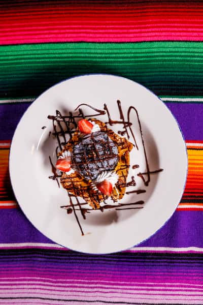 Plated Mexican dessert featuring a chocolate-drizzled pastry topped with whipped cream, strawberries, and a scoop of dark chocolate ice cream, on a festive striped tablecloth.