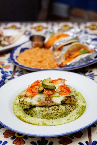 Grilled chicken breast smothered in melted cheese and topped with shrimp and avocado slices, served over cilantro-lime rice and surrounded by a creamy green chile sauce on a blue-rimmed white plate.