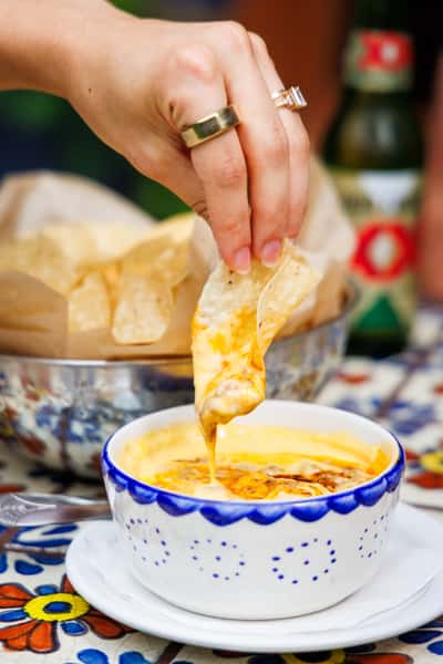 Hand dipping a crispy tortilla chip into a hot bowl of creamy queso dip with swirls of melted cheese and roasted peppers, accompanied by a basket of tortilla chips—ideal for a Tex-Mex appetizer spread.