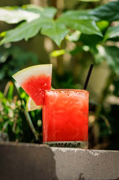 Icy watermelon margarita in a chili-rimmed glass, garnished with a fresh watermelon wedge and lime slice, set against a lush green garden—perfect for summer sipping.