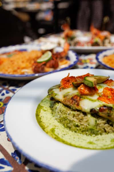 Grilled chicken breast smothered in melted cheese and topped with shrimp and avocado slices, served over cilantro-lime rice and surrounded by a creamy green chile sauce—vibrant Mexican entrée on a Talavera-tiled table.